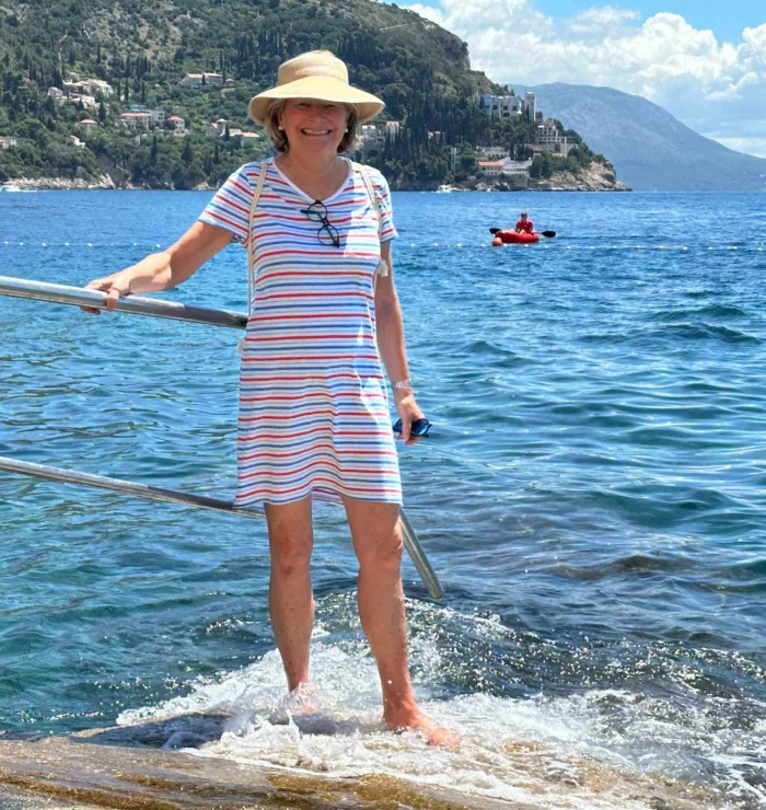 A woman in a striped dress and sun hat stands on a rock by the sea. A person in a red kayak is in the distance. Hills and houses are visible in the background.