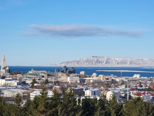 Aerial view of Reykjavik with its colorful buildings, church spire, harbor, and distant snow-capped mountains under a clear blue sky.