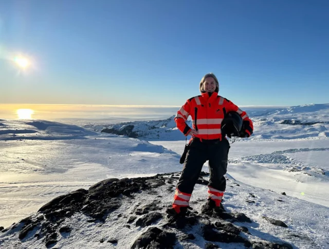 A person in a bright orange and black outfit stands on a snowy mountain under a clear blue sky with the sun shining.