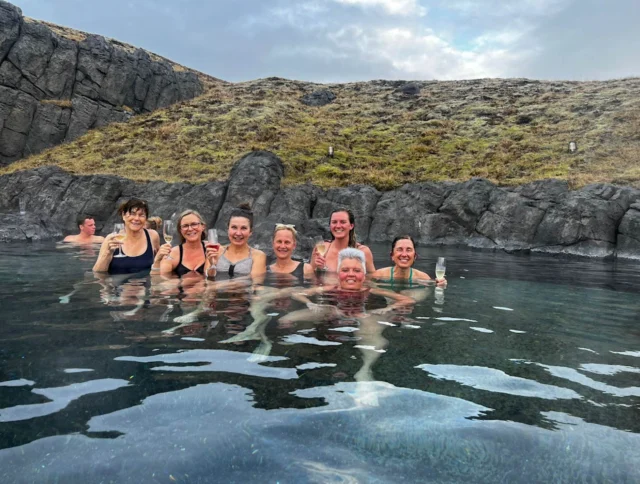 A group of people smiling and holding drinks while relaxing in a natural hot spring surrounded by rocks and grass under a cloudy sky.