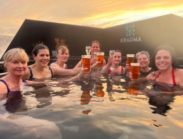 Seven women in swimsuits holding drinks, standing in an outdoor hot spring with a sunset in the background.