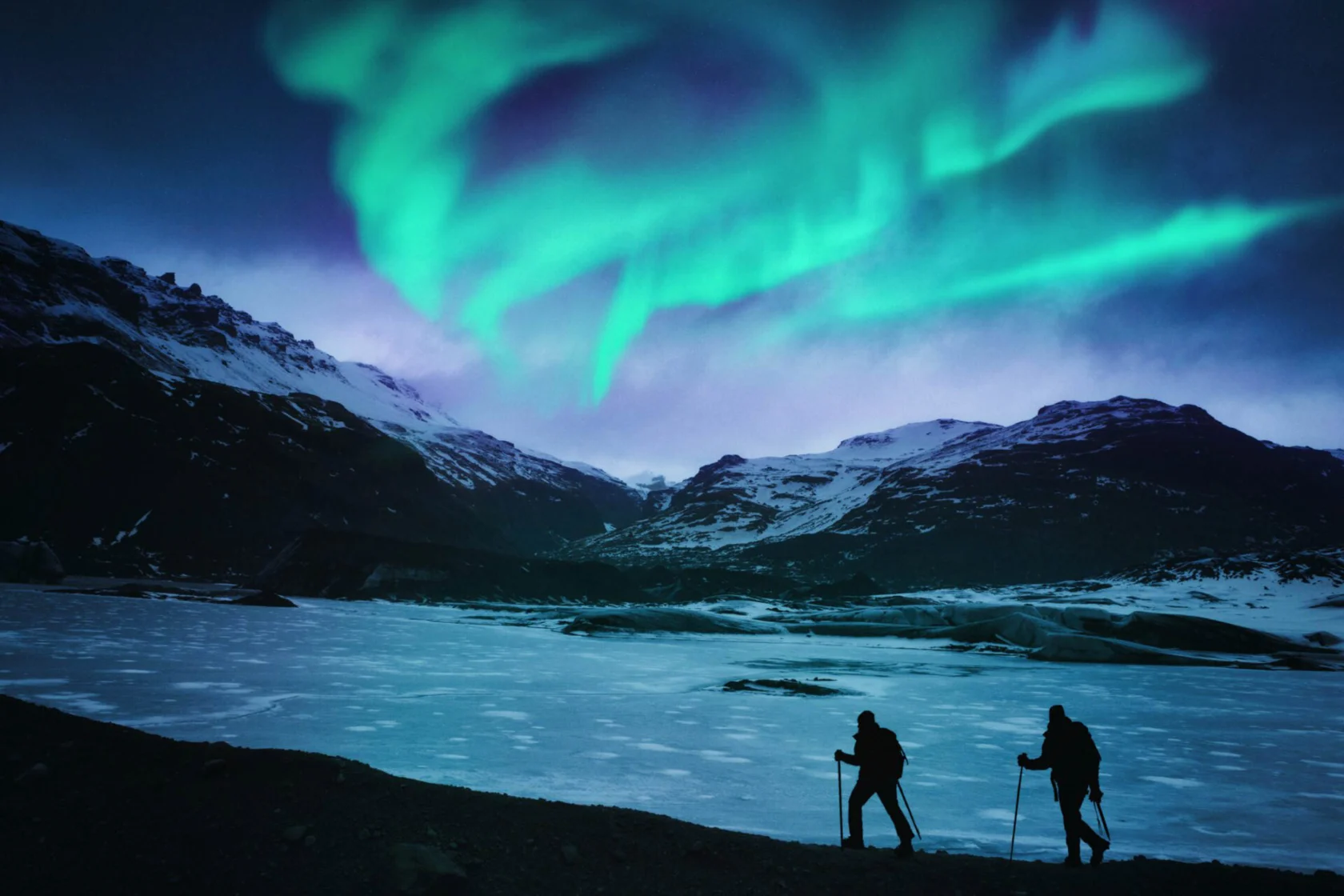 Two people hike with trekking poles under the northern lights, with snow-covered mountains in the background.