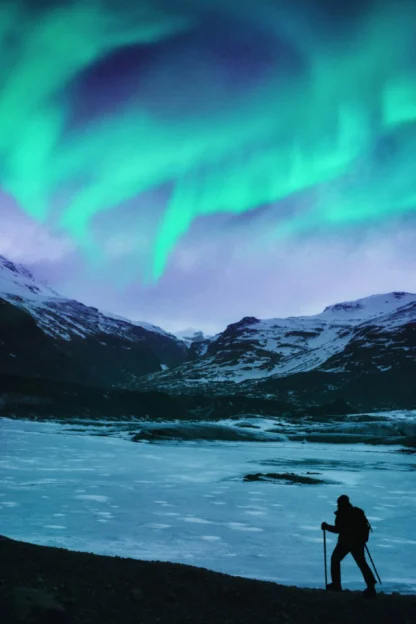 Two people hike with trekking poles under the northern lights, with snow-covered mountains in the background.