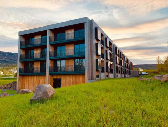 A modern, rectangular building with large windows and wooden accents sits on a grassy landscape under a partly cloudy sky.