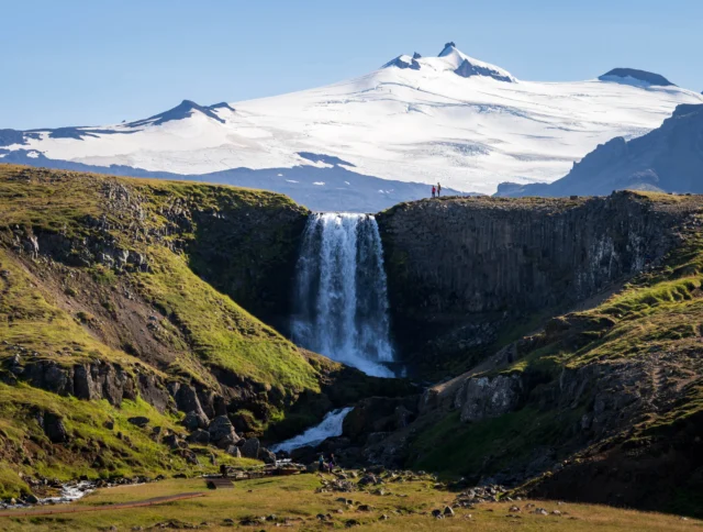 A waterfall cascades over a cliff with a snow-capped mountain in the background, surrounded by lush greenery under a clear blue sky.