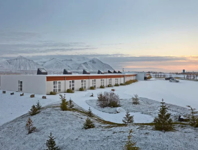 A long building with solar panels on the roof is surrounded by a snowy landscape with distant mountains under a cloudy sky.