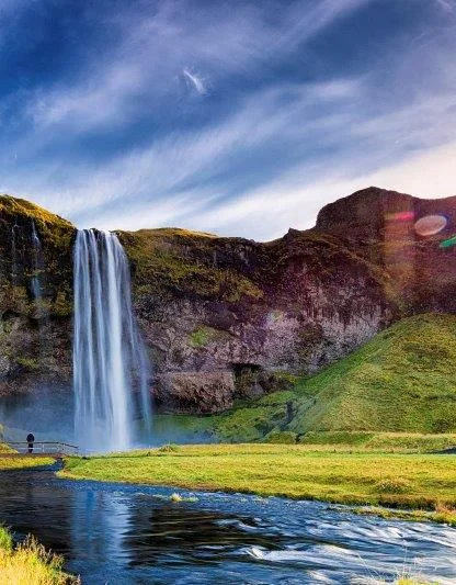 A tall waterfall cascades down a rocky cliff into a river, surrounded by green hills. The sun is setting, casting a bright glow in the sky.