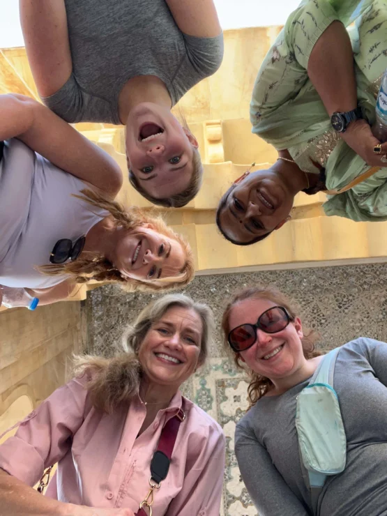 Five people standing in a circle and smiling down at the camera. Three women wear casual tops, and two wear long-sleeved outfits. They are outdoors with a stone wall in the background.