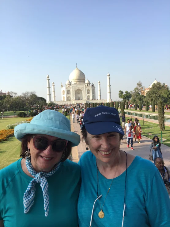 Two women wearing hats and blue clothing stand smiling in front of the Taj Mahal on a sunny day, surrounded by tourists.