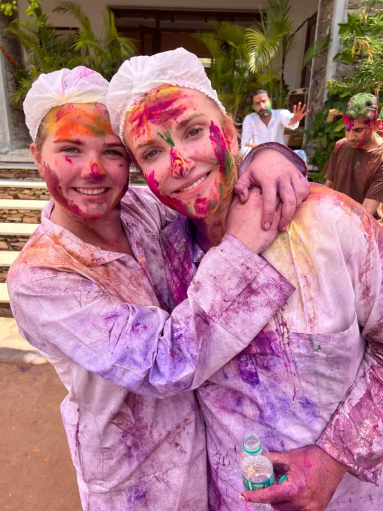 Two people covered in colorful powder smiling and hugging, wearing protective hair caps and white clothing during what appears to be a Holi celebration. People and plants are visible in the background.