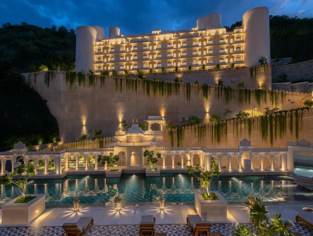 A luxurious, illuminated hotel at night with a grand pool in front, surrounded by ornate architecture and greenery on a hillside.