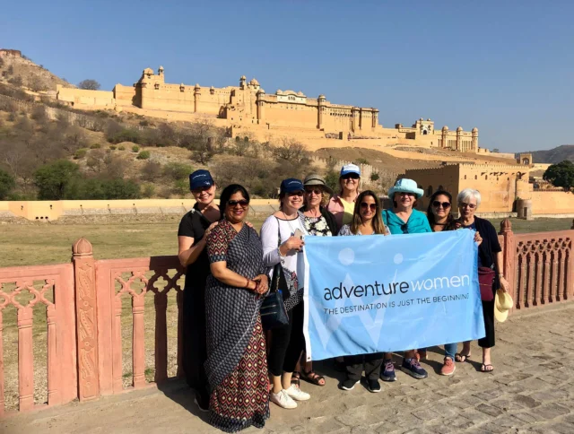 A group of women poses with a banner reading "Adventure Women" in front of a historic fort on a sunny day.