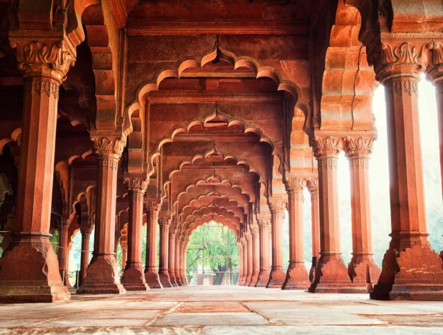 A series of intricately designed red sandstone arches in a historical architectural setting, with sunlight streaming through.