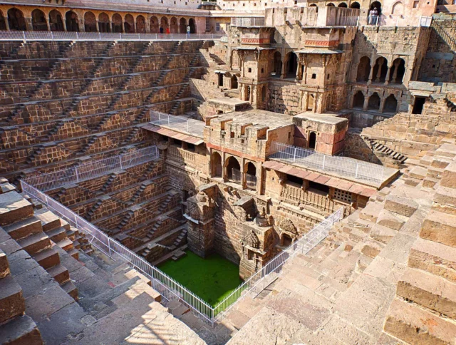 Ancient stepwell with geometric stone steps and platforms leading to green water at the bottom, surrounded by intricate stone architecture.