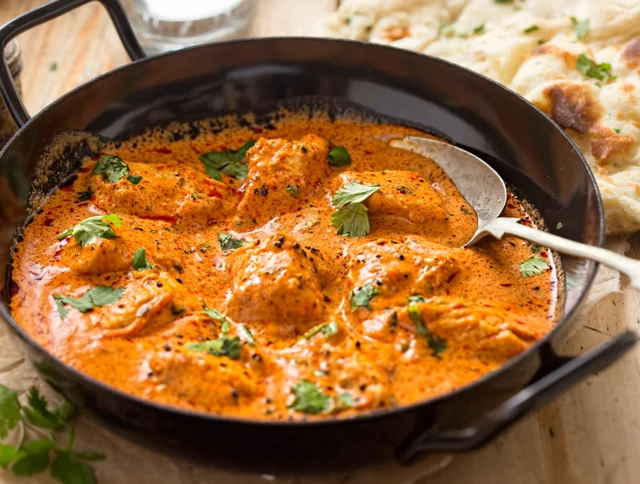 A black pan filled with creamy orange butter chicken, garnished with cilantro. A spoon rests in the dish. Naan bread is visible on the side.