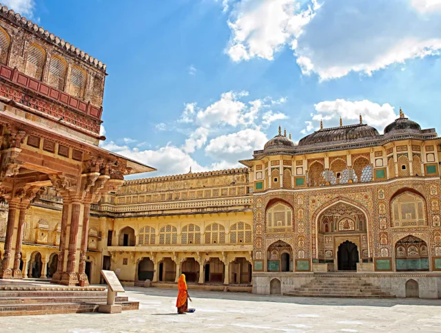 A person in an orange robe walks through a courtyard of ornate, historic buildings under a partly cloudy sky.