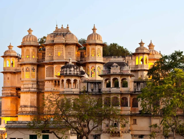 A grand, historic palace with multiple domes and intricate windows, surrounded by trees, illuminated against a blue sky at dusk.