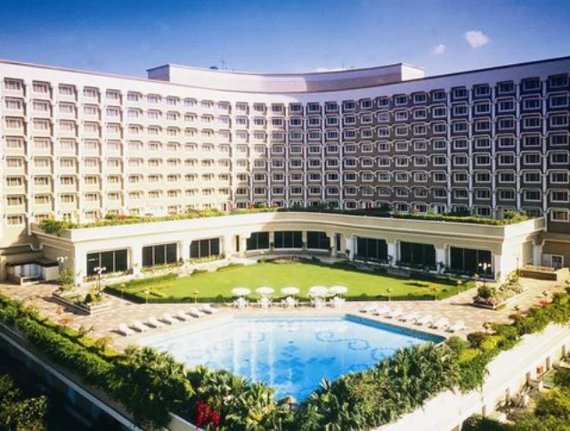 A large, multi-story hotel building with a U-shaped design features a central courtyard with a swimming pool surrounded by greenery and patio furniture under a clear blue sky.