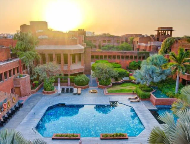 A terracotta brick hotel complex with a central blue pool surrounded by greenery. The sun sets in the background, casting a warm glow over the scene.