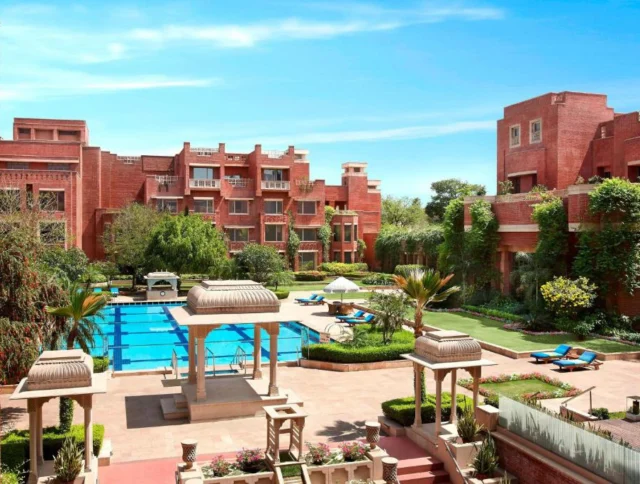 A red brick building surrounds a courtyard with a swimming pool, lush greenery, lounge chairs, and a decorative pavilion under a clear blue sky.