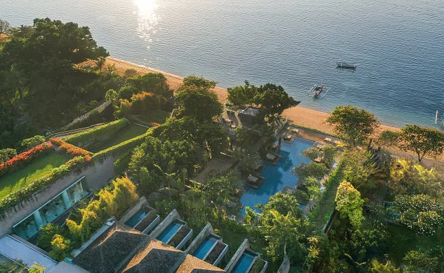 Aerial view of a coastal resort with a pool, lush greenery, and a sandy beach next to a calm sea.