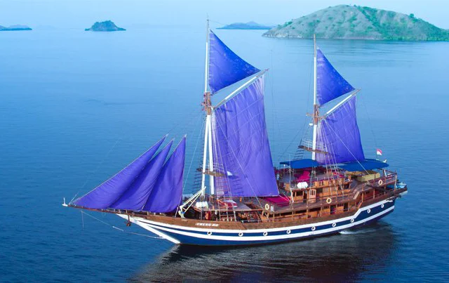 A large traditional sailing ship with bright purple sails glides on calm blue waters, with small islands in the distant background.