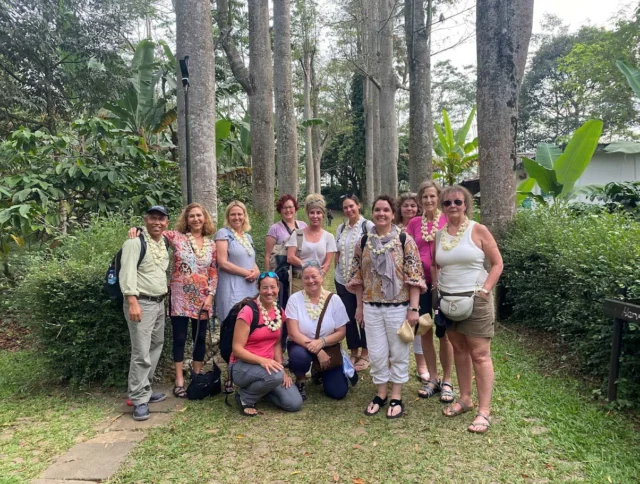 A group of people standing together in a forested area, surrounded by tall trees and greenery. Some wear leis and casual clothing.