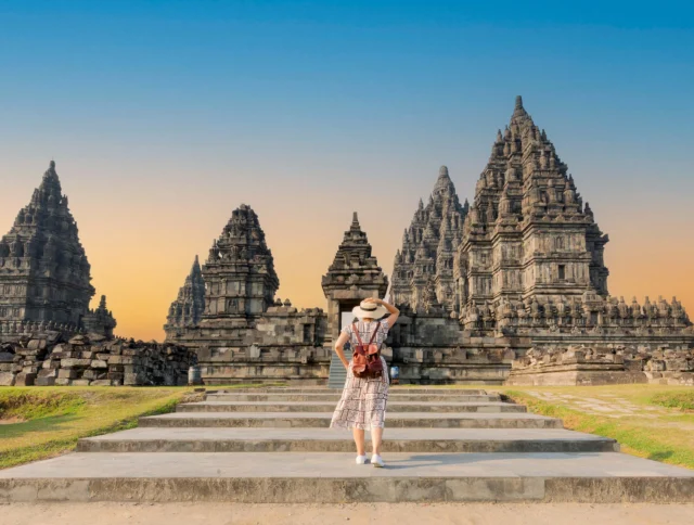 A person in a dress and hat stands in front of ancient stone temples under a clear sky.