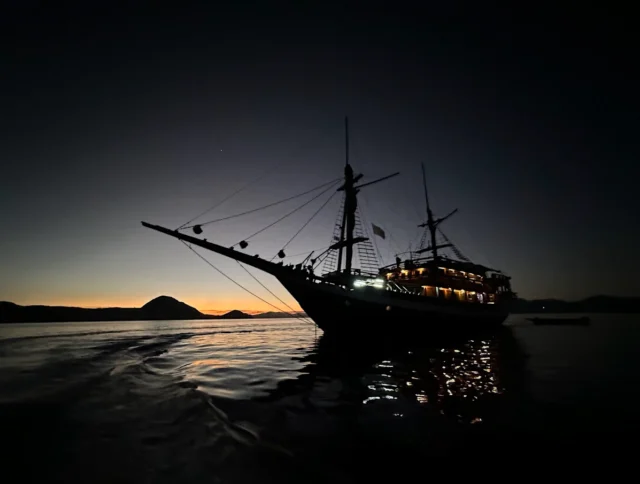 Silhouette of a large sailing ship on calm water at dusk, with a faint orange glow on the horizon and dark mountains in the background.