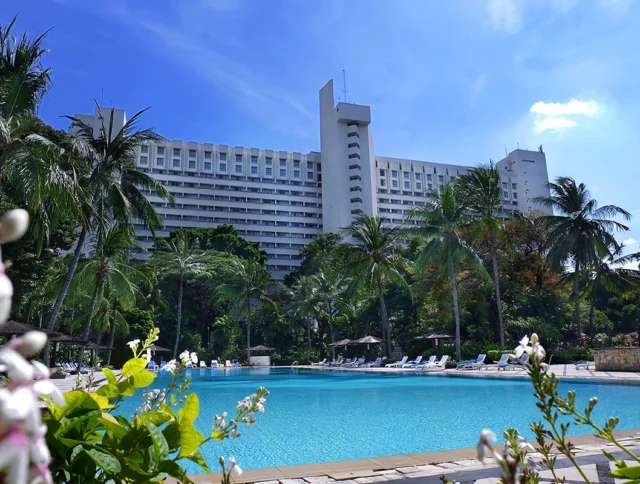 A large hotel with a modern design is situated behind a turquoise swimming pool, surrounded by palm trees and lush greenery under a clear blue sky.