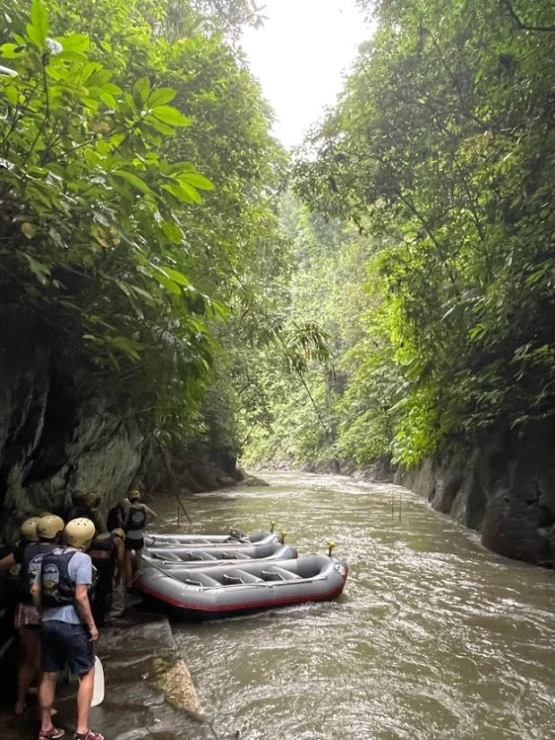 People wearing helmets and life jackets stand beside rafts on a narrow, jungle-lined river.