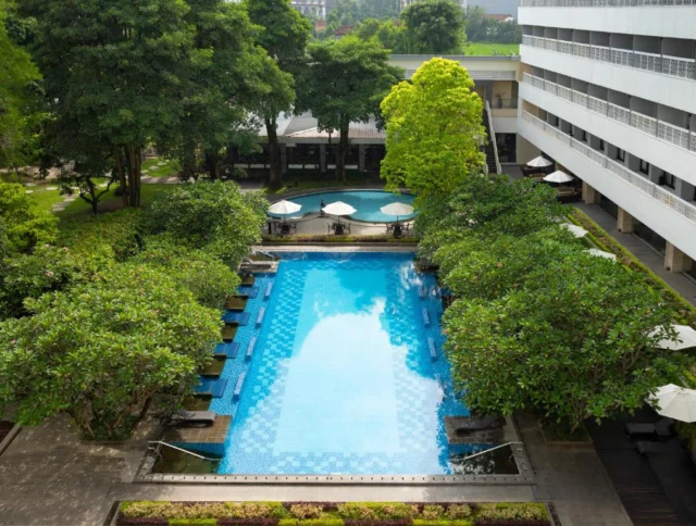 Aerial view of a rectangular outdoor pool surrounded by trees, with lounge chairs and umbrellas along the sides, adjacent to a multi-story building.