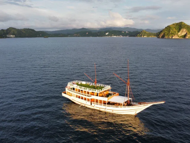 A large wooden boat with two masts floats on calm water near green, hilly islands under a partly cloudy sky.