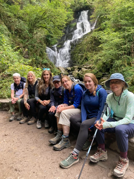 Seven people sitting on a bench in front of a waterfall, surrounded by greenery. They are dressed in outdoor gear and appear to be on a hiking trip.