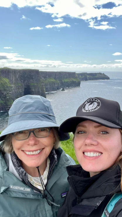 Two people smiling in front of a coastal cliff landscape, with cloudy skies and calm ocean water. Both wear hats and outdoor jackets.