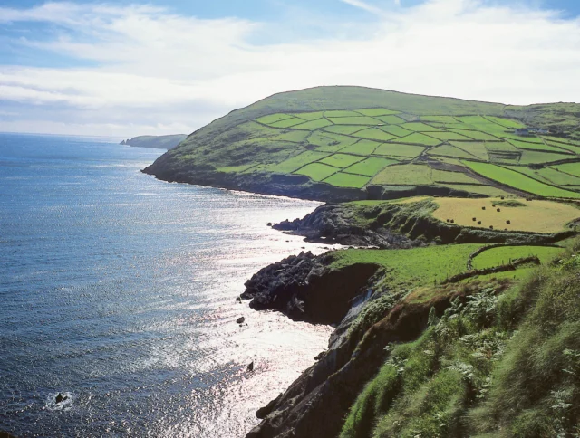 Coastal landscape featuring rolling green hills and patchwork fields alongside a calm blue sea under a partly cloudy sky.