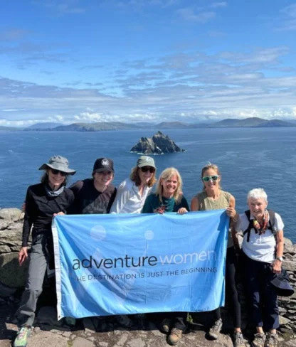 A group of women holding an "Adventure Women" banner stands on a rocky coastal viewpoint with islands in the background.