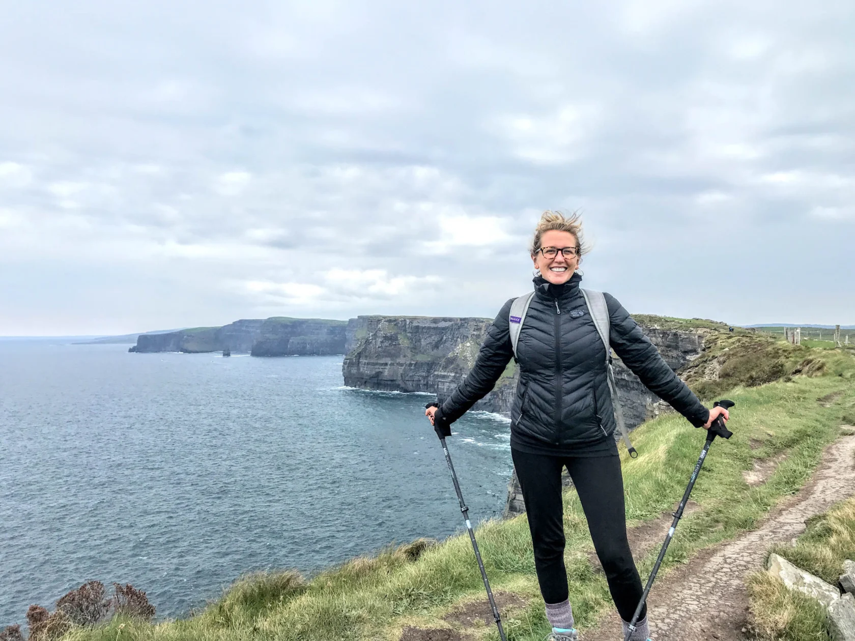 A person stands smiling on a coastal trail holding trekking poles, with cliffs and the sea in the background under a cloudy sky.