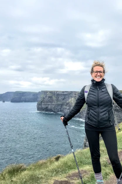 A person stands smiling on a coastal trail holding trekking poles, with cliffs and the sea in the background under a cloudy sky.