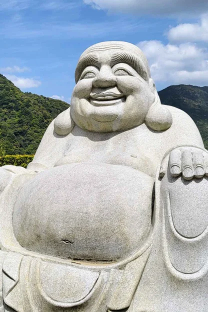 A large stone statue of a seated, smiling figure with a bald head and a round belly in front of a lush, green mountainous landscape under a blue sky.