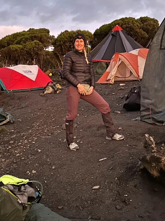 Person standing on a campsite with tents and gear around, wearing a jacket, hat, and gaiters.