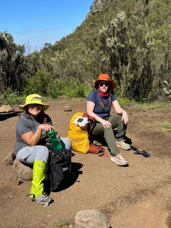 Two people are sitting on a rocky path in a hilly area, wearing hats and outdoor gear, with backpacks and a water bottle nearby.