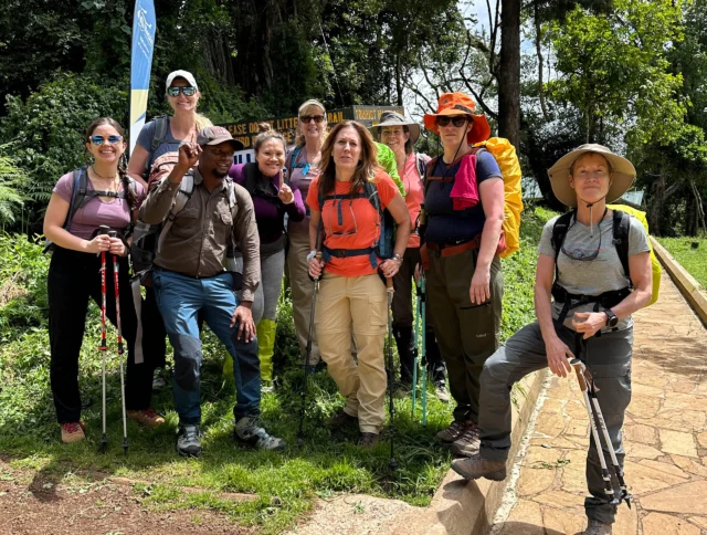 A group of hikers poses for a photo on a path, surrounded by greenery and trees. Some hold trekking poles, and a sign is visible in the background.