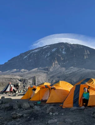 Orange tents set up on a rocky mountain base under a clear blue sky, with a person peeking out from one tent. A large mountain looms in the background.