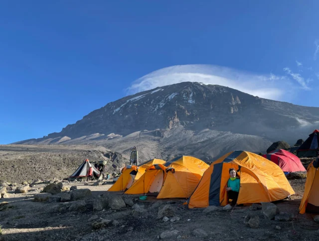 Orange tents set up on a rocky mountain base under a clear blue sky, with a person peeking out from one tent. A large mountain looms in the background.