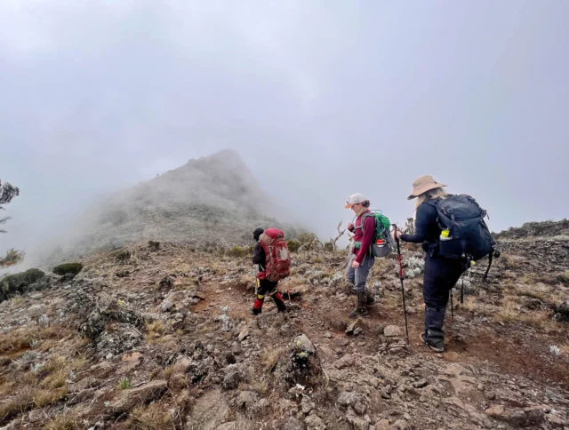 Three hikers in outdoor gear walk on a rocky mountain trail shrouded in mist, surrounded by rugged terrain.