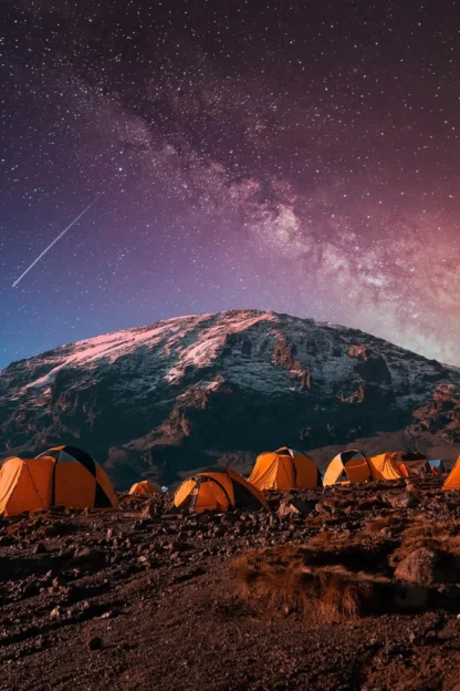 A group of tents on a rocky landscape under a starry night sky with a snow-capped mountain in the background.