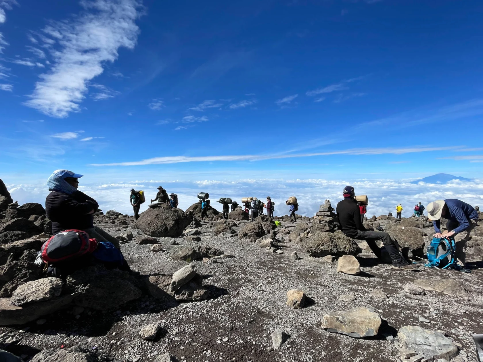 People sitting on rocks at a mountain summit, overlooking clouds and distant peaks under a clear blue sky.