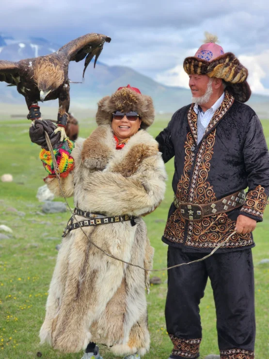 Two people in traditional attire stand on grass in a mountainous landscape. One holds a large bird of prey on their arm.