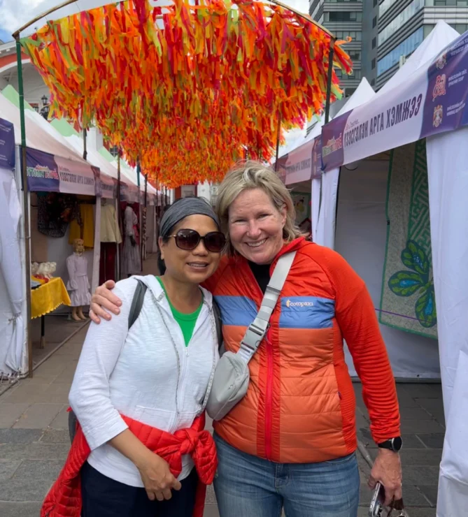 Two people smiling and posing together under a colorful canopy at an outdoor market with tents in the background.